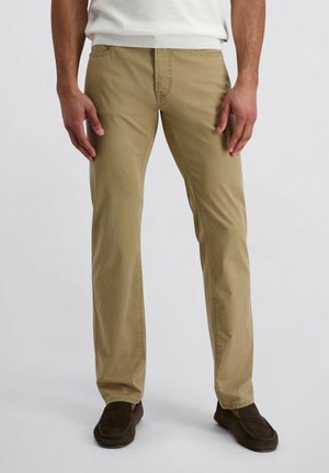 Mężczyzna w jasnobrązowych spodniach chinos i ciemnobrązowych butach typu slip-on stoi na jednolitym tle.