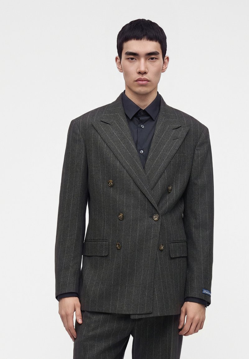 Jeune homme portant une veste de costume croisée gris foncé à fines rayures sur une chemise noire boutonnée, debout devant un fond clair uni.
