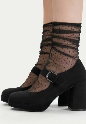 Zapatos Mary Jane de ante negro con tacones de bloque, usados con calcetines tobilleros negros transparentes con lunares sobre piernas desnudas.
