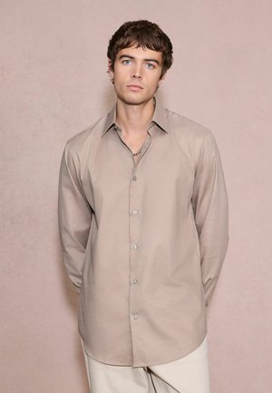 Young man with short brown hair wearing a beige button-up shirt and light pants against a plain beige background.