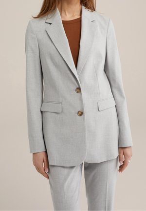 Kvinde iført lysegrå skræddersyet blazer med to knapper over brun ribbet top, kombineret med matchende grå bukser, stående mod ensfarvet baggrund.