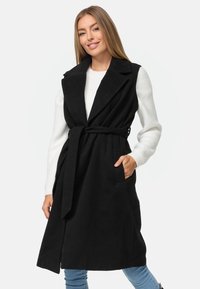 Schwarze Longline-Weste aus einem weichen Stoff, mit einem ausgeschnittenen Kragen, tailliert mit Gürtel und seitlichen Taschen. Über einem weißen Pullover und blauen Jeans getragen.