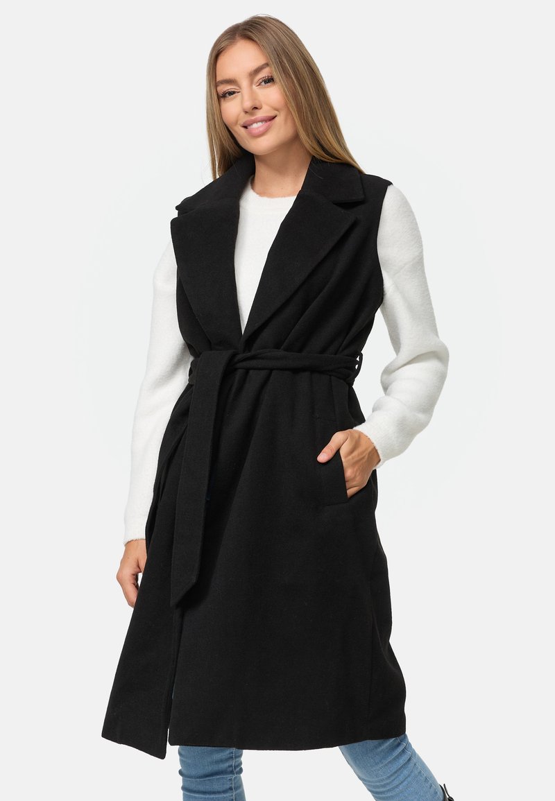 Schwarze Longline-Weste aus einem weichen Stoff, mit einem ausgeschnittenen Kragen, tailliert mit Gürtel und seitlichen Taschen. Über einem weißen Pullover und blauen Jeans getragen.