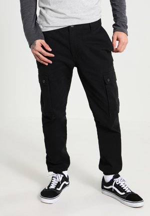 Man draagt zwarte cargobroek met knoopzakken aan de zijkant, zwart-witte sneakers en een grijs langarmshirt met een lichtgrijze voorkant.