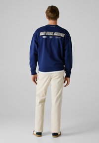 Sweatshirt bleu marine avec le logo Red Bull Racing au dos, poignets et ourlet côtelés, associé à un pantalon cargo crème et des chaussures décontractées.