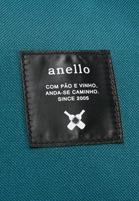 Étiquette en tissu noire sur textile bleu sarcelle avec texte blanc "anello," Expression portugaise sur le pain et le vin, et deux icônes de bouteilles de vin croisées.
