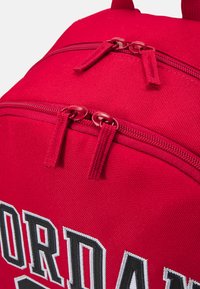 Sac à dos en tissu rouge avec deux compartiments zippés, arborant un logo brodé noir et blanc bien en vue à l'avant. Matériau texturé.