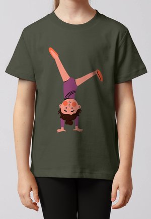 Kind trägt dunkelgrünes Hemd mit Illustration eines Mädchens im lila Trikot, das einen Handstand mit gespreizten Beinen macht.
