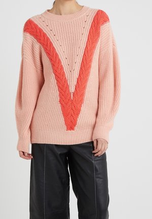 Person trägt einen pinken, grob gestrickten Pullover mit einem leuchtend orangefarbenen, V-förmigen Zopfmuster und schwarze, weit geschnittene Hosen.