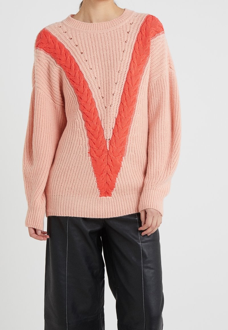 Personne portant un pull en maille épaisse rose avec un motif en forme de V tressé orange vif et un pantalon large noir.