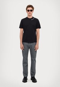 T-shirt nera in cotone con maniche corte, jeans in denim grigi e scarpe nere. Il modello indossa occhiali da sole scuri e si trova di fronte a uno sfondo chiaro.