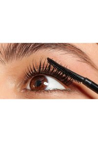 Un pinceau de mascara noir est appliqué sur des cils longs et recourbés, renforçant leur volume sur un œil brun avec une texture de peau lisse et claire.