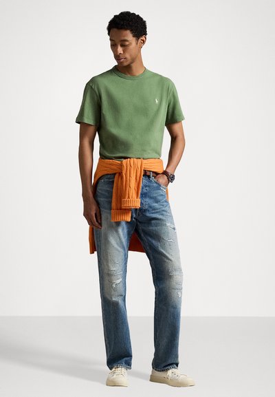 Grön bomullströja med rund hals, i kombination med en orange tröja knuten i midjan. Bärs med slitna blå jeans och ljusa skor.