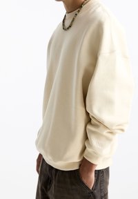 Beżowa bluza z opadającymi ramionami, ściągaczami i subtelnym detalem logo na rękawie. Wykonana z miękkiego, teksturowanego materiału.