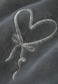 Tissu en polaire gris avec des broderies blanches représentant un cœur et un motif de ruban. La texture est douce et moelleuse, mettant en valeur les coutures détaillées.