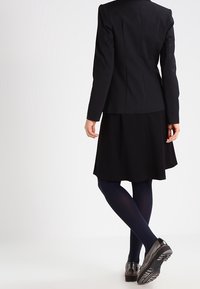 Svart skräddarsydd blazerkavaj med vadderade axlar, matchad med en knälång svart kjol. Bärs med marinblå strumpbyxor och glänsande svarta skor.