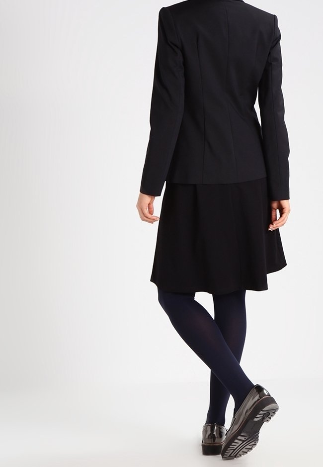 Svart skräddarsydd blazerkavaj med vadderade axlar, matchad med en knälång svart kjol. Bärs med marinblå strumpbyxor och glänsande svarta skor.
