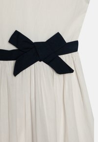 Vestido color crema con un cinturón de tela azul marino, que presenta un gran lazo. La falda tiene pliegues, añadiendo textura y volumen al diseño.
