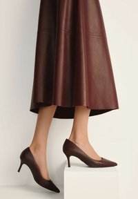 Nogi w burgundowych skórzanych szpilkach o spiczastym nosku i pasującej skórzanej spódnicy midi, prawą nogę uniesioną na białym blokowaniu.