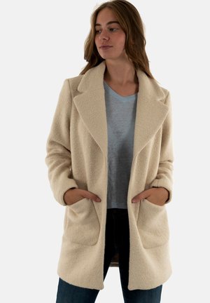 Abrigo color crema hecho de una tela suave y texturizada. Presenta grandes solapas, bolsillos laterales y un corte relajado. Se lleva sobre una blusa de color azul claro.