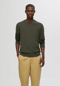 Mörkgrön stickad tröja med rund hals, långa ärmar och ribbad mudd, i kombination med ljus khaki-byxor. Slät textur, avslappnad passform.