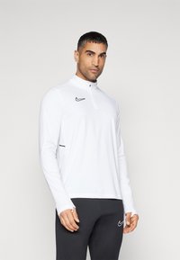 Weißer Langarm-Pullover mit einem Viertelreißverschluss, versehen mit einem schwarzen Nike-Logo auf der Brust und schwarzen Akzenten an den Seiten. Glatte Stofftextur.