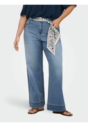 Kvinne iført vide lyseblå jeans med et mønstret skjerfbelte og brune sandaler, står med en hånd i lomma.