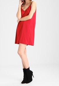 Robe rouge sans manches avec décolleté en V, coupe ample, en tissu lisse. Associée à des bottines noires à bout pointu.