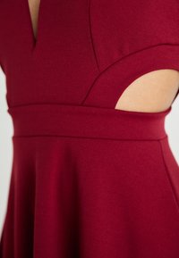 Bordowa sukienka z dekoltem w serek, krótkimi rękawami i wycięciami po bokach. Wykonana z gładkiej tkaniny, z dopasowaną talią i rozkloszowanym dołem.