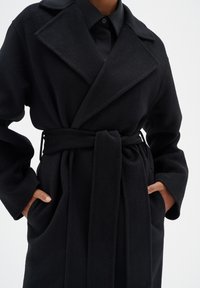 Manteau en polaire noir, oversize, avec un large col, une ceinture à la taille et des poches latérales. Texture lisse et design minimaliste.
