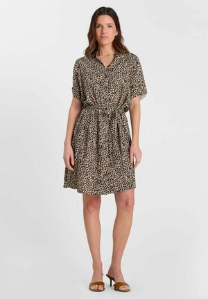 Femme debout portant une robe boutonnée à manches courtes imprimée léopard avec une ceinture à la taille et des sandales à talons ouvertes marron.