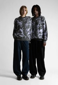 Dwie osoby stojące obok siebie w dopasowanych szarych swetrach z dużymi fioletowymi czaszkami, w luźnych spodniach i czarnych butach.