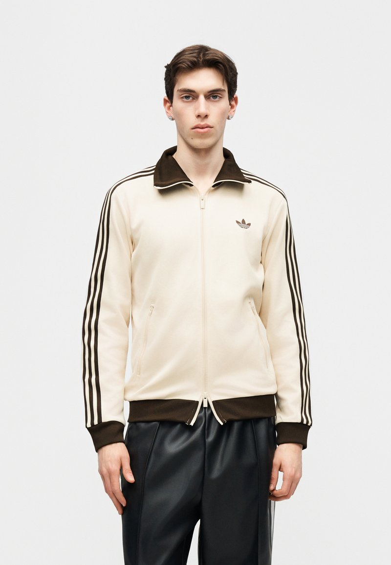 Joven con una chaqueta deportiva Adidas beige y marrón con tres franjas en las mangas y pantalones de cuero negros, de pie frente a un fondo liso.