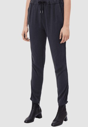Mujer con pantalones negros con cordón y botines negros de tacón, de pie frente a un fondo liso.