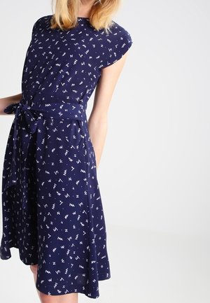 Femme portant une robe bleu marine mi-longue avec un motif dispersé de lettres blanches et une ceinture nouée à la taille, debout devant un fond clair uni.
