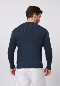 Navy ribbed sweater with long sleeves and a crew neckline. Textured pattern with subtle vertical lines. Worn with white pants.