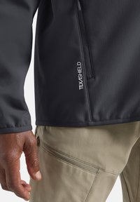 Veste softshell noire en tissu texturé avec une poche zippée, associée à un pantalon beige clair avec une poche zippée sur le côté.