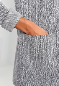 Šedá látka s motivem herringbone, texturovaný povrch; zahrnuje kontrastní světle šedý manžet a boční kapsu viditelnou na pravé straně.