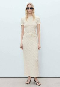 Robe midi crème texturée avec des manches courtes, silhouette ajustée, détail de nœud sur le côté, associée à des lunettes de soleil noires et des sandales à lanières.