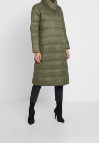 Abrigo de plumas largo en verde oliva con un collar alto, diseño acolchado horizontal y bolsillos laterales, combinado con jeans negros y botas de tacón alto.