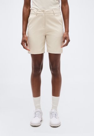 Άτομο που φοράει μπεζ σορτς προσαρμοσμένης ραφής, λευκές κάλτσες και λευκά παπούτσια με κορδόνια, στέκεται μπροστά σε ένα απλό φωτεινό φόντο.