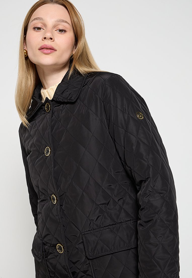 Veste noire matelassée avec un col, ornée de boutons en métal doré et d'un patch logo sur la manche gauche. Le matériau présente une texture lisse et brillante.