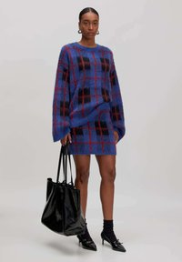 Frau in einem blauen und roten karierten, flauschigen Pullover und passendem Rock, die eine große schwarze Tasche hält, schwarze Socken und spitze Schuhe trägt.