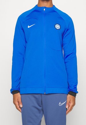 Mężczyzna noszący niebieską sportową kurtkę na zamek błyskawiczny z logo Nike i herbem klubu piłkarskiego Inter Mediolan, w połączeniu z niebieskimi spodniami Nike.