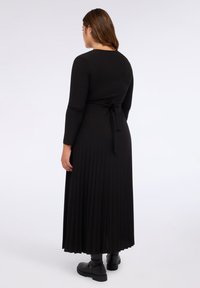 Rochie maxi plisată neagră, cu mâneci lungi și detaliu cu siret la talie. Material neted, ajunge până la gleznă. Purtați cu ghete negre până la gleznă.
