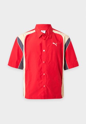 Mand iført en rød, beige og sort kortærmet skjorte med knapper fra Puma og lyseblå jeans med Puma-logoer.