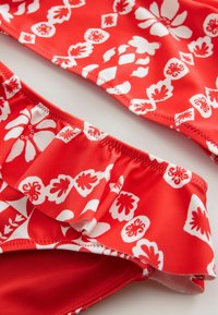 Czerwona tkanina bikini z białymi kwiatowymi i geometrycznymi wzorami; posiada falbany i gładką teksturę; zaokrąglone krawędzie i bezszwowy design.
