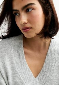Pull gris à col en V en tissu doux et texturé ; design ajusté avec un col côtelé. Cheveux foncés, aux épaules, avec un maquillage naturel.