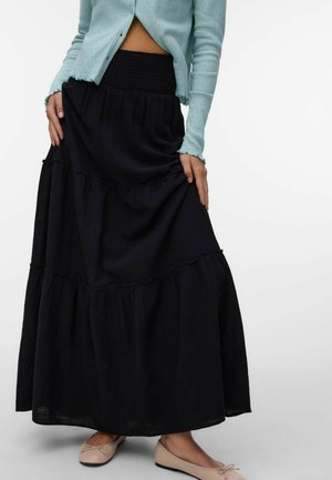 Femme portant une longue jupe noire à volants, un cardigan bleu clair court et des ballerines beiges, tenant la jupe à deux mains.