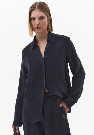 Femme blonde aux cheveux raides portant une chemise ample bleu marine foncé à boutons et un pantalon assorti, tenant un petit sac à main sombre.